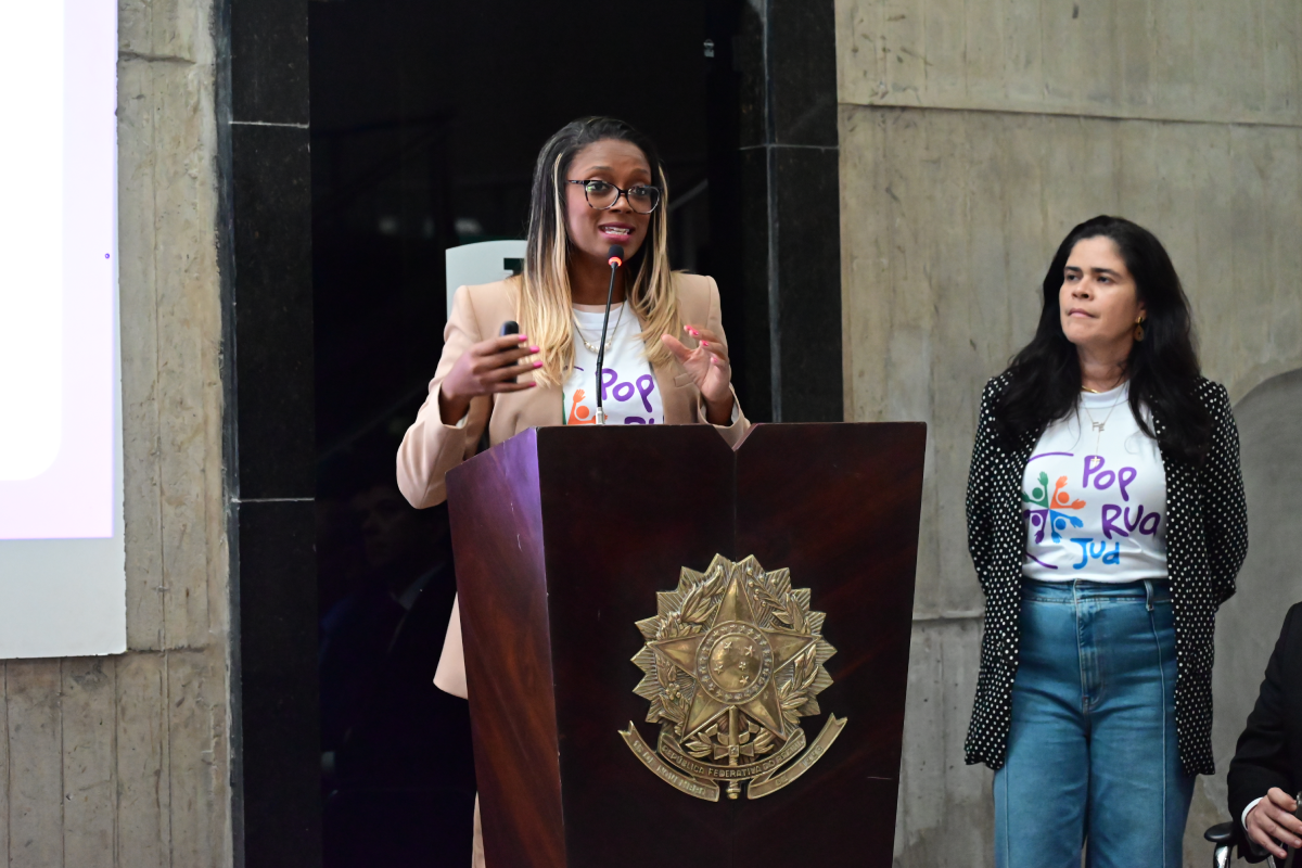Uma mulher negra de cabelos loiros e óculos discursa em um púlpito de madeira com o brasão da República, vestindo blazer bege sobre a camiseta do "PopRuaJud". Ela gesticula com as mãos enquanto fala ao microfone, sob o olhar de outra mulher à direita que veste blazer de poá e calça jeans. O ambiente tem paredes de concreto aparente e um painel de projeção à esquerda.